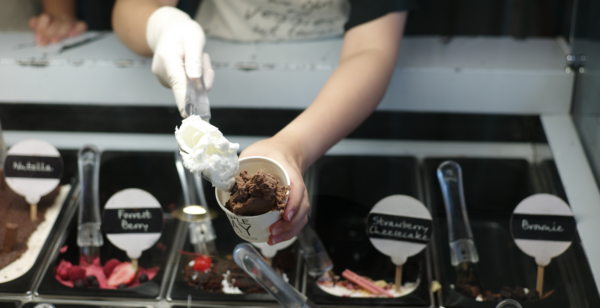 A person at a scoop shop putting a scoop of gluten-free vanilla ice cream into a bowl of chocolate ice cream.
